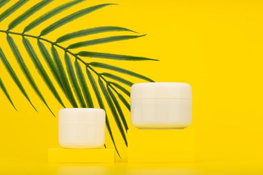 Two white jars with cosmetic products for skin care on yellow podium against yellow background with palm leaf. Concept of summer skin care or organic cosmetics with natural ingredients