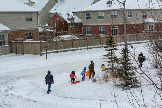 Children and their parents walk down the snow-covered sidewalk to the slide, carrying a plastic sled