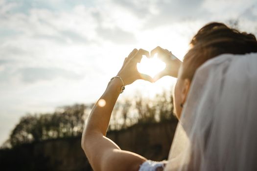 bride with a veil on her head looks at the sun through her heart-shaped hands