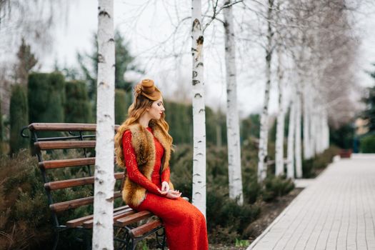 young girl with red hair in a bright red dress on a bench in an empty park among the birches