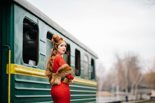 young girl with red hair in a bright red dress near an old green passenger car