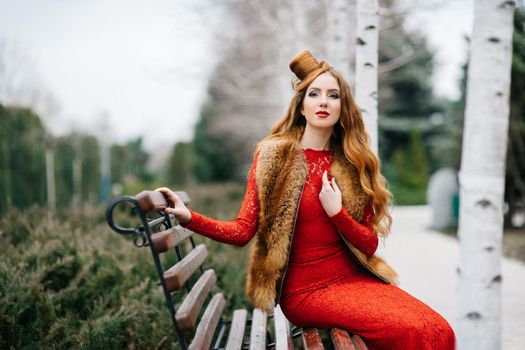 young girl with red hair in a bright red dress on a bench in an empty park among the birches