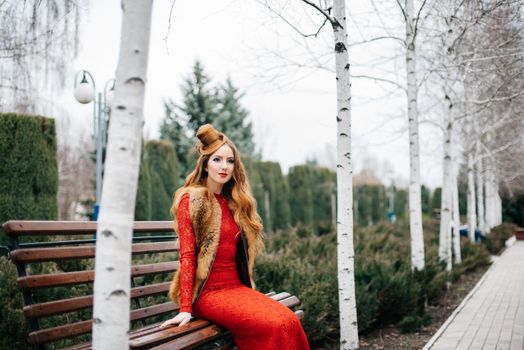 young girl with red hair in a bright red dress on a bench in an empty park among the birches