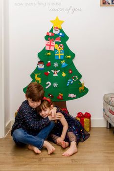 A cute, brown-haired, blue-eyed baby girl and a cute, red-haired, blue-eyed boy smartly dressed hugging in the floor in front of a Christmas tree