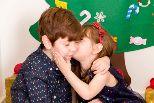 A cute, brown-haired, blue-eyed baby girl and a cute, red-haired, blue-eyed boy smartly dressed kissing in front of a Christmas tree