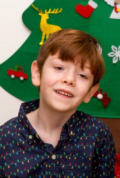 A  cute, red-haired, blue-eyed boy smartly dressed smiling in front of a Christmas tree