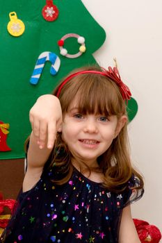 A  cute, brown-haired, blue-eyed baby smartly dressed smiling in front of a Christmas tree with her hand up