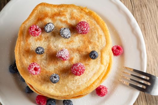 Stack of pancakes with fresh berries on a white plate on the table, top view.