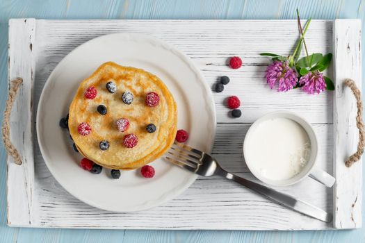 Stack of pancakes with fresh berries on a white plate a cup of milk and wildflowers on the table, top view.