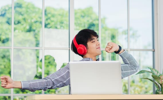 Asian young business man smile listening music in red headphone at home office. Happy businessman sitting on desk and relax wear wireless head phones enjoy listen music and work with laptop computer