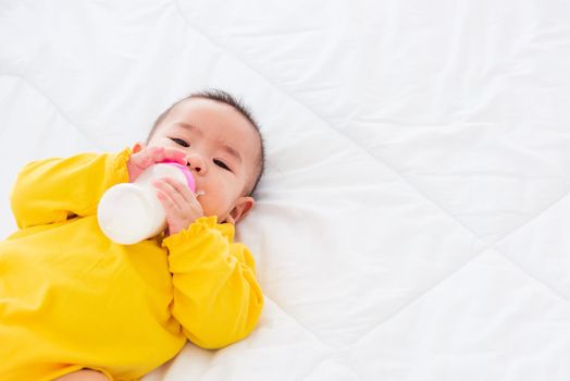 Asian beautiful little baby girl wearing a yellow dress eat milk sleeping feeding lying on the white bed, infant holding a bottle of milk, baby food concept