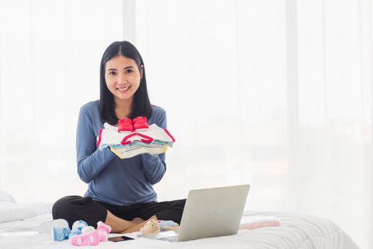 Asian mother shopping online for her baby clothes on laptop computer and tablet on the bed she makes purchase new baby clothes for an unborn baby preparing for the new child