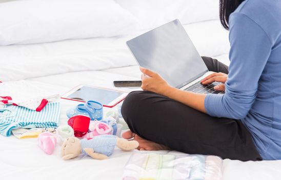 Asian mother shopping online for her baby clothes on laptop computer and tablet on the bed she makes purchase new baby clothes for an unborn baby preparing for the new child