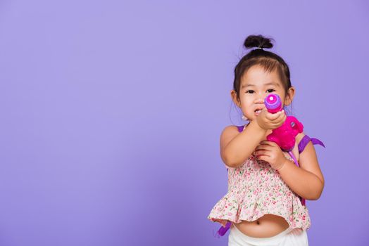 Thai child funny hold toy water pistol and smile, Happy Asian little girl holding plastic water gun, studio shot isolated on purple background, Thailand Songkran festival day national culture concept