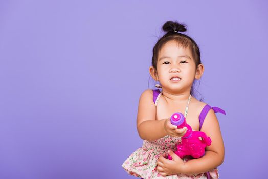 Thai child funny hold toy water pistol and smile, Happy Asian little girl holding plastic water gun, studio shot isolated on purple background, Thailand Songkran festival day national culture concept