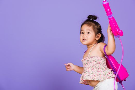 Thai child funny hold toy water pistol and smile, Happy Asian little girl holding plastic water gun, studio shot isolated on purple background, Thailand Songkran festival day national culture concept