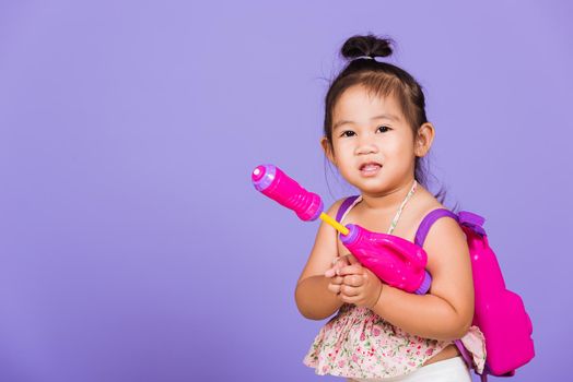 Thai child funny hold toy water pistol and smile, Happy Asian little girl holding plastic water gun, studio shot isolated on purple background, Thailand Songkran festival day national culture concept