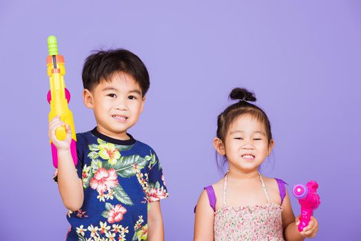 Two Happy Asian little boy and girl holding plastic water gun, Thai children funny hold toy water pistol and smile, studio shot isolated on purple background, Thailand Songkran festival day culture.