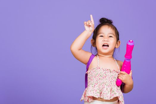 Happy Asian little girl holding plastic water gun, Thai child funny hold toy water pistol and smile, studio shot isolated on purple background, Thailand Songkran festival day national culture concept