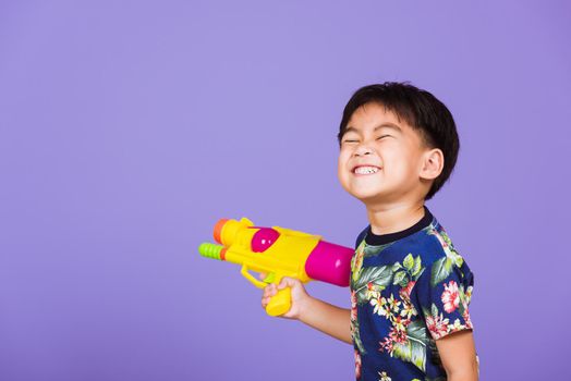 Thai kid funny hold toy water pistol and smiling, Happy Asian little boy holding plastic water gun, studio shot isolated on purple background, Thailand Songkran festival day national culture concept