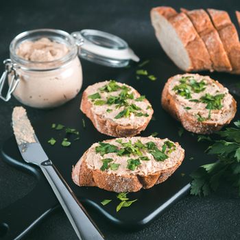 Close up view of slice bread with homemade turkey pate and fresh green parsley on black kutting board over black cement background,