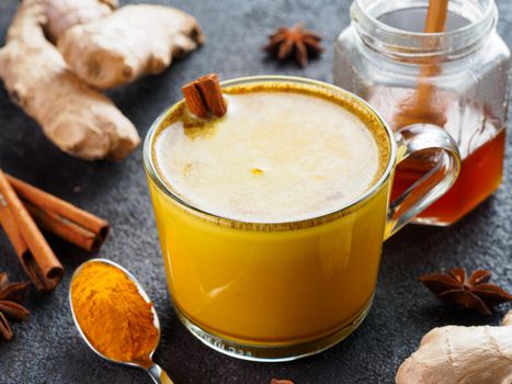 Healthy drink golden turmeric latte in glass cup.Gold milk with turmeric,ginger root,cinnamon sticks,dried turmeric powder and honey over black cement background.Detox turmeric tea and ingredients.
