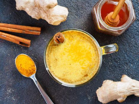 Healthy drink golden turmeric latte in glass cup.Gold milk with turmeric,ginger root,cinnamon sticks,turmeric powder and honey on black background.Detox turmeric tea and ingredients.Top view flat-lay.