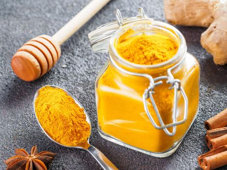 Dried turmeric powder in glass jar and spoon on black cement background. Ingredients for golden milk, turmeric latte or detox drink - honey dipper, ginger root, cinnamon sticks and star anise