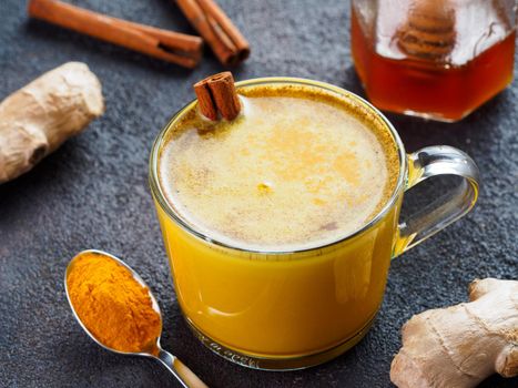 Healthy drink golden turmeric latte in glass cup.Gold milk with turmeric,ginger root,cinnamon sticks,dried turmeric powder and honey over black cement background.Detox turmeric tea and ingredients.