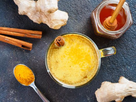 Healthy drink golden turmeric latte in glass cup.Gold milk with turmeric,ginger root,cinnamon sticks,turmeric powder and honey on black background.Detox turmeric tea and ingredients.Top view flat-lay.