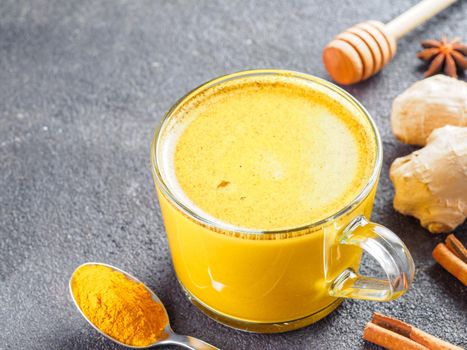 Healthy drink golden turmeric latte in glass cup.Gold milk with turmeric,ginger root,cinnamon sticks,turmeric powder and honey over black cement background.Detox turmeric tea and ingredients.Copyspace