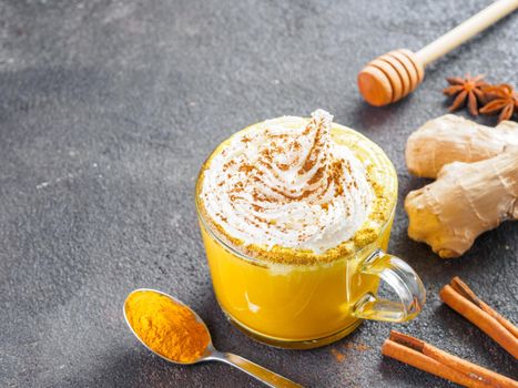 Healthy drink golden turmeric latte with whipped cream in glass cup. Gold milk,ginger root,cinnamon sticks,turmeric powder and honey over black background.Detox turmeric tea and ingredients.Copyspace