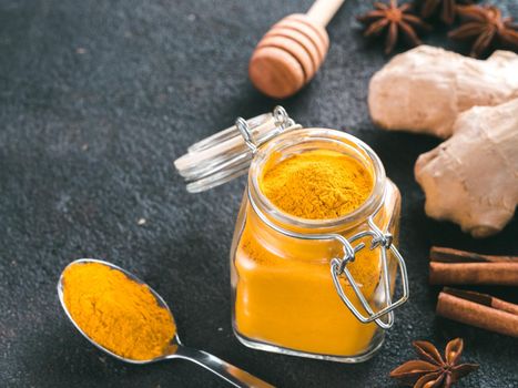 Dried turmeric powder in glass jar and spoon on black cement background.Ingredients for golden milk,turmeric latte,detox drink.Honey dipper, ginger root, cinnamon sticks, star anise.Copy space. Toned