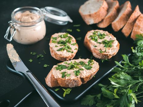 Close up view of slice bread with homemade turkey pate and fresh green parsley on black kutting board over black cement background,