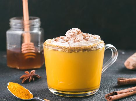 Healthy drink golden turmeric latte with whipped cream in glass cup. Gold milk,ginger root,cinnamon,turmeric powder and honey on black background.Detox turmeric tea and ingredients.Copy space.Toned