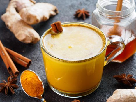 Healthy drink golden turmeric latte in glass cup.Gold milk with turmeric,ginger root,cinnamon sticks,dried turmeric powder and honey over black cement background.Detox turmeric tea and ingredients.
