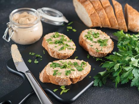 Close up view of slice bread with homemade turkey pate and fresh green parsley on black kutting board over black cement background,