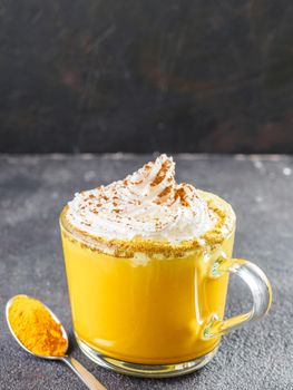 Healthy drink golden turmeric latte with whipped cream in glass cup. Gold milk, and dried turmeric powderin spoon on black cement background. Detox turmeric tea. Copy space. Vertical.