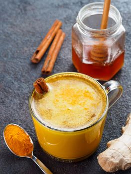 Healthy drink golden turmeric latte in glass cup.Gold milk with turmeric,ginger root,cinnamon sticks,turmeric powder and honey over black cement background.Detox turmeric tea and ingredients.Copyspace