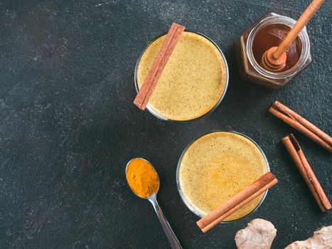 Healthy drink golden turmeric latte in glass. Gold milk,ginger root,cinnamon sticks,turmeric powder and honey on black cement background.Detox turmeric tea and ingredients. Copy space. Top view. Toned
