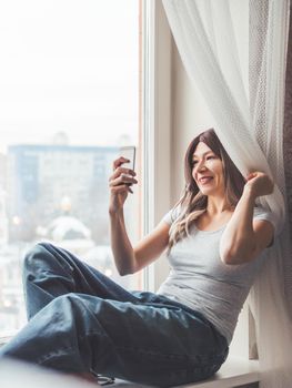 Woman with curly hair poses for selfie on windowsill. Pretty woman makes self-portrait on smartphone. Winter morning at home.