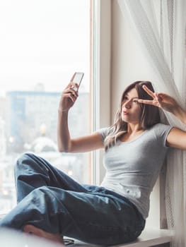 Woman with curly hair poses for selfie on windowsill. Pretty woman makes self-portrait on smartphone. Winter morning at home.