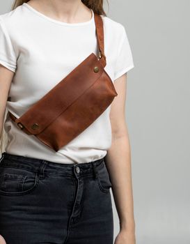 Girl in a white blouse with a leather brown handmade bag over her shoulder. Designer brown banana bag. Woman in a studio. Comfortable small bag for walking