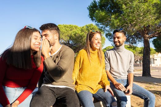 Two young beautiful heterosexual couple laughing each other. Happy millennial group of people having fun outdoor. Man speaks in the ear of her girlfriend sitting on a wall of the city square