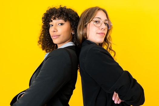 Confident multiracial business women back to back with crossed arms looking at camera isolated on yellow background for copy space. Female success on work and better life concept