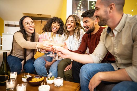 Multiracial young happy friends group at home sitting on the sofa toasting with champagne - Mixed race millennial laughing people celebrating clinking with wine glasses and table laden with snacks