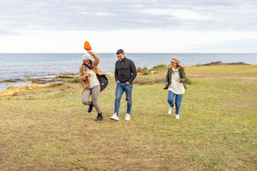 Three mixed-race carefree friends running outdoor joking each other - Millennial people having fun on a meadow near sea smiling in winter vacation enjoying nature in ocean resort wearing clothing