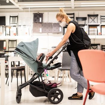 Young mom with newborn in stroller shopping at retail furniture and home accessories store wearing protective medical face mask to prevent spreading of corona virus. New normal during covid epidemic.