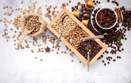 Green and brown decaf unroasted and dark roasted coffee beans in wooden box with scoops setup on white concrete background.