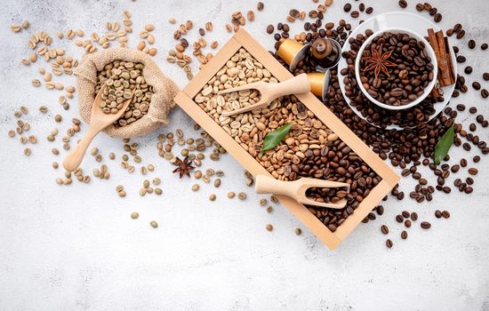 Green and brown decaf unroasted and dark roasted coffee beans in wooden box with scoops setup on white concrete background.